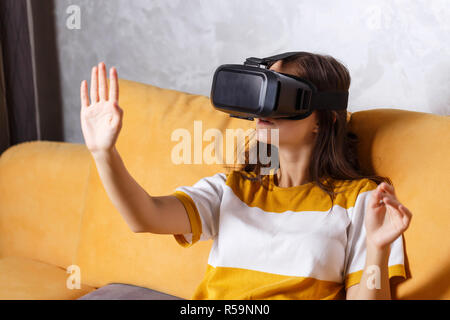 Cute long-haired girl in pullover wearing a virtual reality headset while sitting on the yellow sofa in the light living room, future technology concept Stock Photo