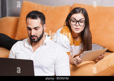 Pretty long-haired girl in pullover wearing glasses laying with digital tablet on the yellow sofa in the light living room and her bearded multicultural boyfriend in white shirt working on the laptop, girl peeking over shoulder in the laptop, modern technology, communication concept Stock Photo