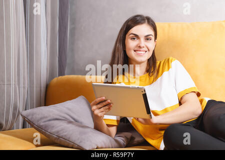 Happy long haired brunette girl laying on the yellow sofa in the light living room and holding digital tablet, modern technology, communication concept Stock Photo