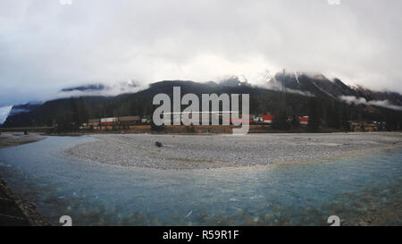 Long cargo train passing through mountains behind small river with azure water during rainy day Stock Photo