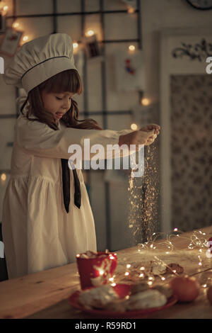 Child girl in a cook cap sprinkles flour on the table on background of garland from light bulbs. Stock Photo