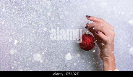 Woman holding red glittering Christmas ball in hand over silver background with snow, light bokeh. Banner, copy space. New year holiday celebration co Stock Photo