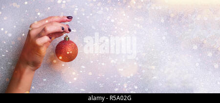 Woman holding red glittering Christmas ball in hand over silver background with snow, light bokeh. Banner, copy space. New year holiday celebration co Stock Photo