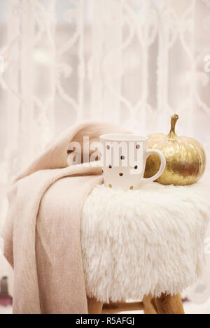 Hot chocolate, a cup of cappuccino on a fur chair in front of a large window with a white sheers curtain. Warm scarf and golden pumpkin around. Cozy w Stock Photo