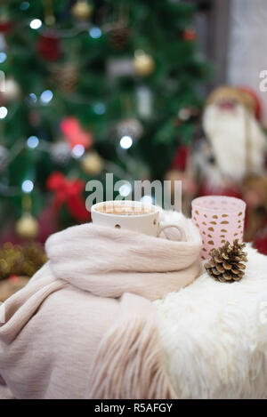 Christmas time. Hot chocolate, a cup of cappuccino on a fur chair in front of a large Christmas tree with balls and lights. Warm scarf, cones around. Stock Photo