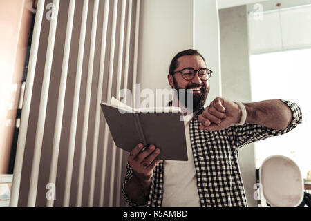 So busy. Cheerful nice man looking at the time while holding his notebook Stock Photo