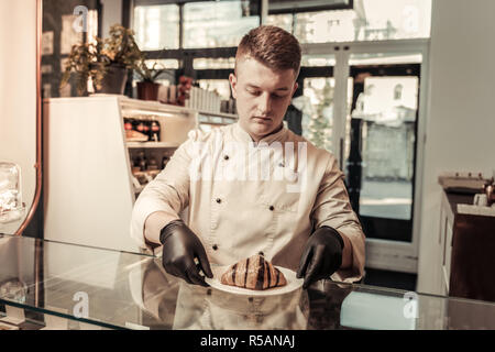 Best bakery. Nice professional baker holding a plate with a croissant while putting it on the display Stock Photo