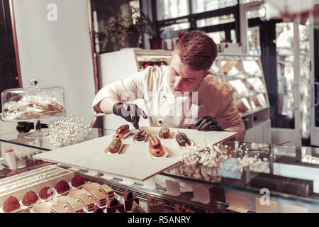 Sweet cakes. Nice professional confectioner looking at the tasty eclairs while decorating them Stock Photo