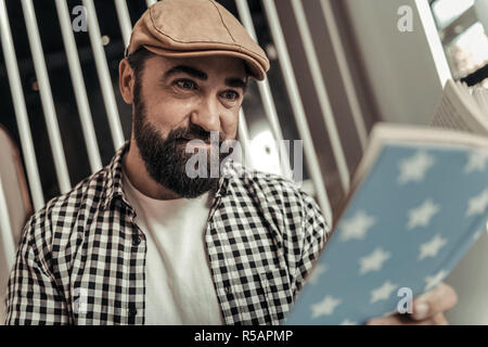 Interesting story. Joyful nice bearded man reading a book while sitting in the cafeteria Stock Photo