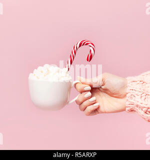 Female hands in knitted sweater holding cup of marshmallows and Christmas candy cane on pink background Flat Lay copy space Winter traditional food Fe Stock Photo