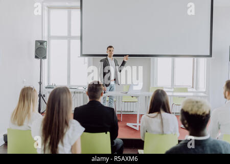 Business coach. Rear view of man gesturing with hand while standing against defocused group of people sitting at the chairs in front of him Stock Photo
