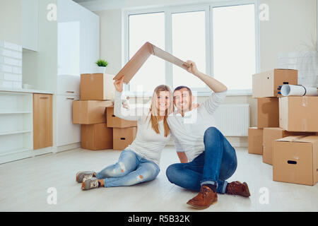 Happy young couple sitting on floor under piece of carton in new apartment with boxes on background smiling at camera Stock Photo