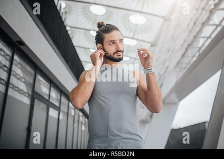 Picture of well-built and handsom bearded young man looks to right. He puts headphones in ears. Guy walks on stadium. He is alone Stock Photo