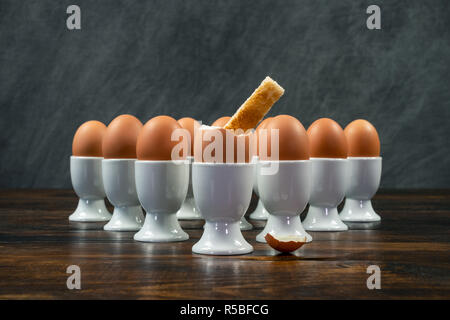 Toast soldier dipped into one boiled egg of a group of eggs in white egg cups on a wooden table Stock Photo