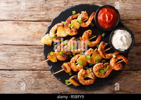 Fresh kebabs made from shrimps and pineapple slices decorated with green onions served with sauces close-up on a slate plate on a wooden table. Horizo Stock Photo