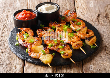 Grilled seafood kebabs from shrimp and pineapple slices with sauces close-up on a slate plate on a wooden table. horizontal Stock Photo