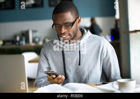 African American millennial student look at smartphone studying in cafe, focused black young guy text using cellphone sitting in coffesshop working at Stock Photo