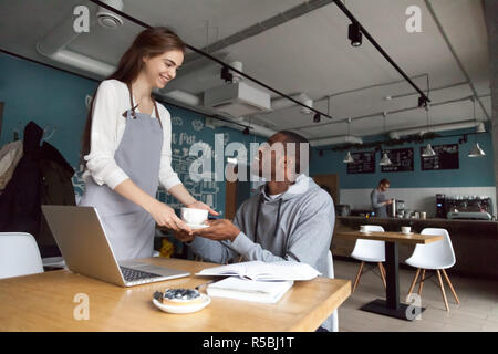 Smiling young waitress serve coffee to African American cafe visitor studying or working at table, pretty coffeehouse female worker give cappuccino or Stock Photo