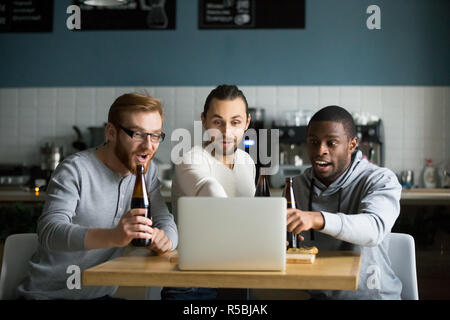 Diverse friends hang out in cafe drinking beer, watching online game at laptop, multiethnic males cheering football team enjoying match sitting in pub Stock Photo