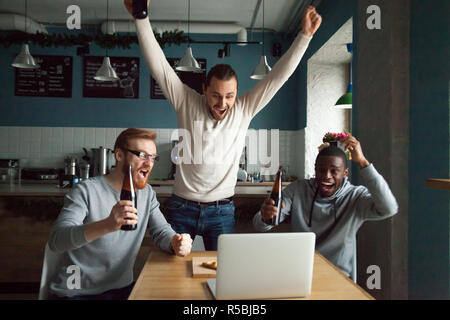 Excited millennial guys celebrating victory watching football match on laptop in pub, diverse males scream supporting sports team scoring goal, drinki Stock Photo