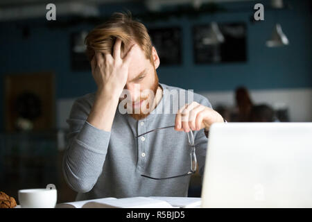 Exhausted red haired student tired of studying too much sitting with laptop and books in cafe, annoyed millennial man suffer from headache working on Stock Photo
