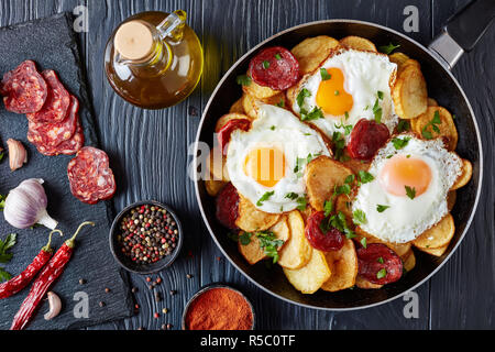 Traditional spanish huevos rotos - fried eggs with potatoes, pork sausages chorizo in a skillet on a black wooden table with ingredients, horizontal v Stock Photo