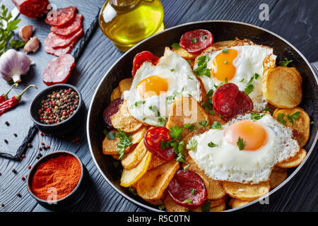 close-up of Traditional spanish huevos rotos - fried eggs with potatoes, pork sausages chorizo in a skillet on a black wooden table with ingredients,  Stock Photo