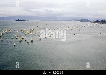 View facing southeast of sailboats passing through San Francisco Bay during the Pacific Inter-Club Yacht Association (PICYA) Opening Day of The Bay Parade, in San Francisco, California, April 30, 1955. In the distance at right is the San Francisco-Oakland Bay Bridge connecting to Treasure Island in the Bay. At left middle ground is the prison island of Alcatraz. () Stock Photo