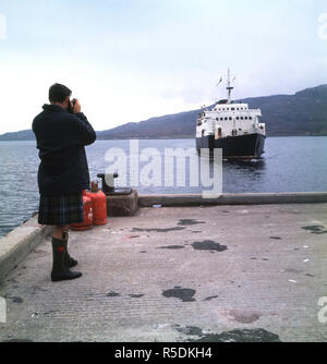 1960s, scottish man wearing a kilt taking a photo of a passenger ferry coming into the harbour of the Isle of Lochmaddy, Outer Hebrides, Western Isles, Scotland. Stock Photo