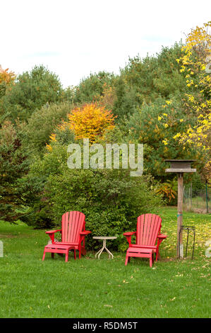 Two red muskoka chairs sit empty in lush green backyard Stock Photo