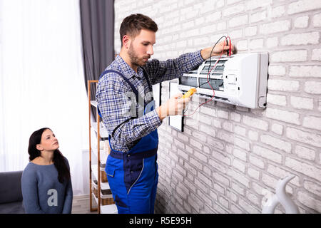 Young Woman Looking At Technician Examining Air Conditioner With Digital Multimeter Stock Photo