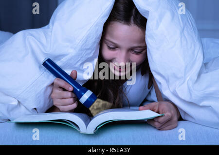 Girl Under White Blanket Using Flashlight While Reading Book At Night Stock Photo