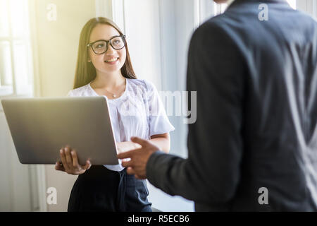 Happy couple of young business partners working in modern office. Two coworkers working on laptop while standing near window. Stock Photo