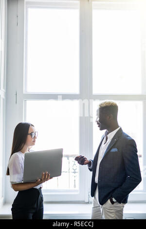 Pretty young smiling woman pointing at laptop while standing near her happy afro business man in front of window in office. Stock Photo