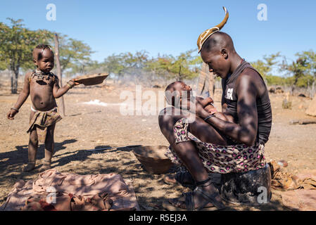 Young Himba boy with a traditional young Himba hairstyle holding a baby on his lap while a child passing by is looking at them. Stock Photo