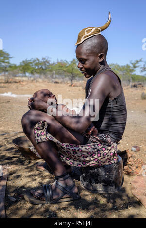 Young Himba boy with a traditional young Himba hairstyle holding a baby on his lap smiling at him. Stock Photo