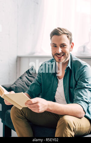 laughing young man holding book and looking at camera on sofa at home Stock Photo