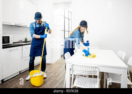Man and woman as a professional cleaners in uniform washing floor and wiping furniture in the white kitchen. Cleaning service concept Stock Photo
