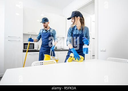 Man and woman as a professional cleaners in uniform washing floor and wiping furniture in the white kitchen. Cleaning service concept Stock Photo
