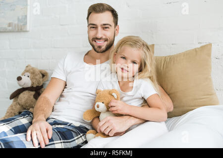happy father and cute little daughter with teddy bear sitting on bed and smiling at camera Stock Photo