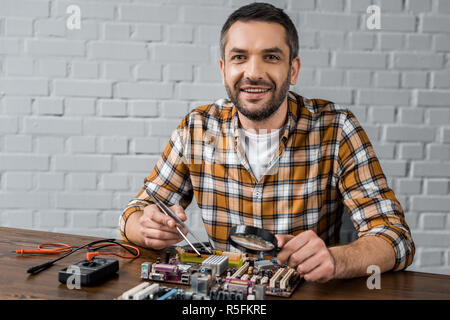 handsome smiling computer engineer with tweezers and magnifying glass repairing motherboard Stock Photo