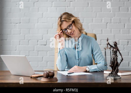 beautiful female lawyer in eyeglasses smiling at camera while sitting at workplace Stock Photo