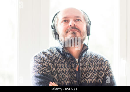 Smiling middle age man listening to music with headphones while standing next to window at home, head and shoulders, music concept Stock Photo