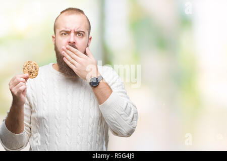 Young hipster man eating chocolate chips cookie over isolated background cover mouth with hand shocked with shame for mistake, expression of fear, sca Stock Photo