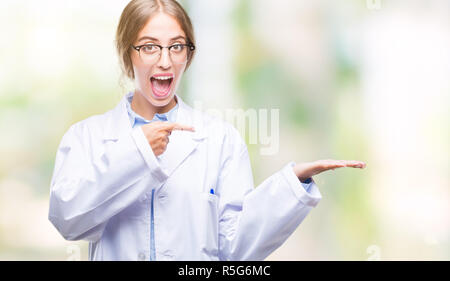 Beautiful young blonde doctor woman wearing white coat over isolated background amazed and smiling to the camera while presenting with hand and pointi Stock Photo