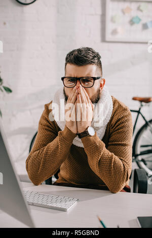 sick businessman in knitted scarf sitting at office desk, coughing and covering mouth with hands Stock Photo