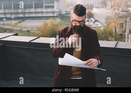 pensive adult male architect in glasses holding blueprint and working on project on rooftop Stock Photo