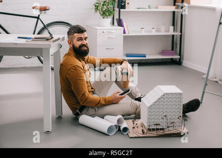 smiling bearded adult male architect using smartphone, holding coffee to go and working on blueprints in office Stock Photo