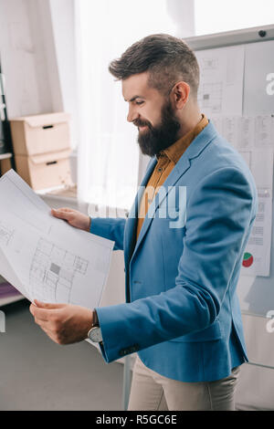 side view of smiling adult male architect in blue formal wear holding blueprint, using flip chart and working on project in office Stock Photo