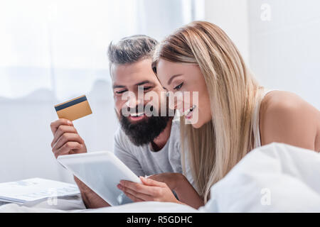 Attractive couple with digital tablet and credit card lying in bright bedroom Stock Photo